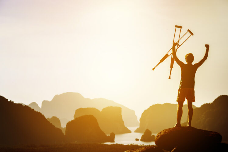 Man with crutches stands in winner pose with rised hands on mountains and islands background. Space for text